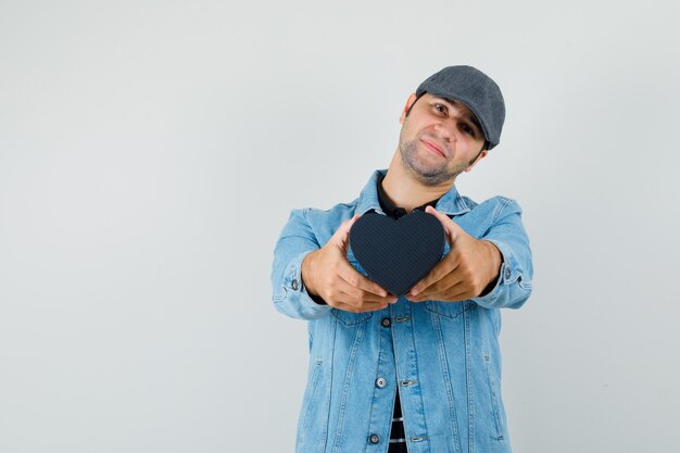 Young man in jacket,cap showing heart-shaped gift and looking lovable space for text