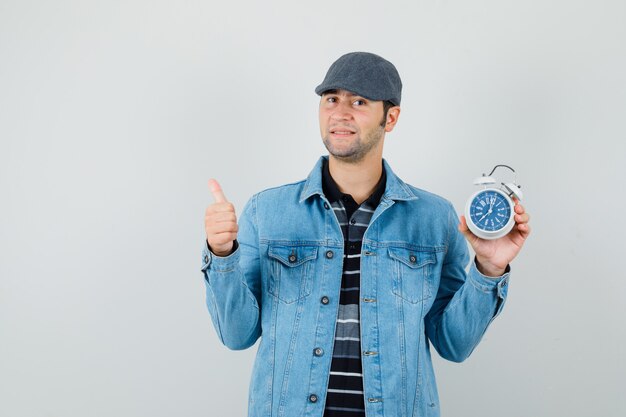 Young man in jacket,cap showing thumb up while holding clock and looking pleased