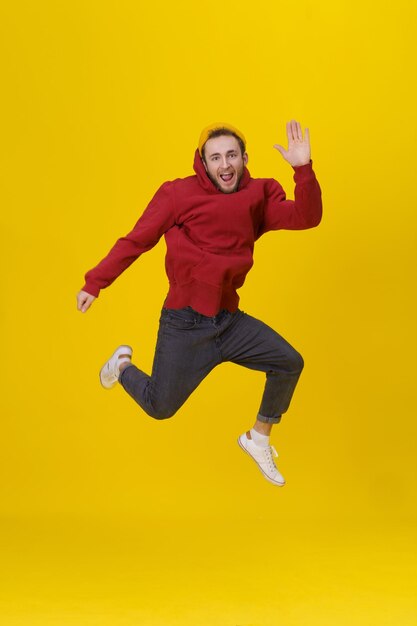 Молодой человек в радости прыгает высоко в повседневной красной толстовке с капюшоном и джинсах, изолированных на желтом