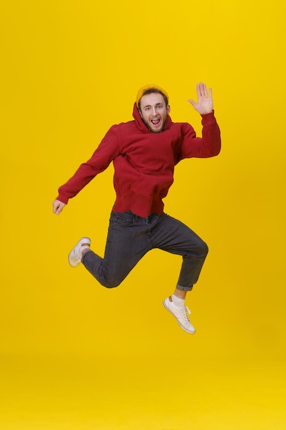 Young man in joy jumping high wearing casual red hoodie and jeans isolated on yellow Humorous hipster guy in jump