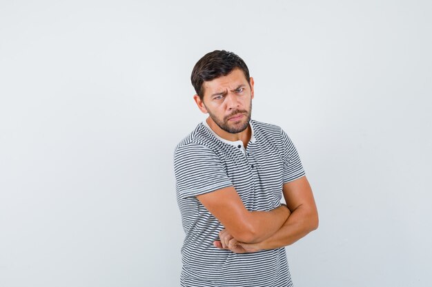 Young man keeping arms folded in t-shirt and looking uncertain , front view.