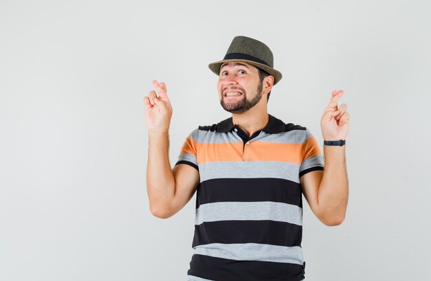 Young man keeping fingers crossed in t-shirt, hat and looking cheerful. front view.