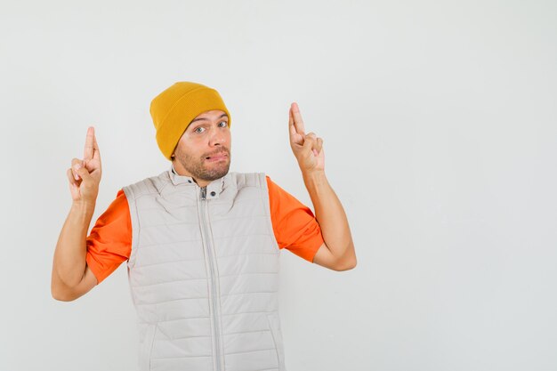 Young man keeping fingers crossed in t-shirt, jacket, hat and looking confused. front view.