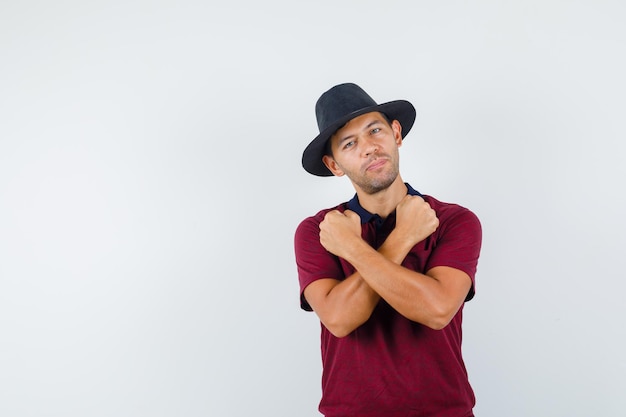 Young man keeping fists crossed in t-shirt, hat and looking confident. front view.