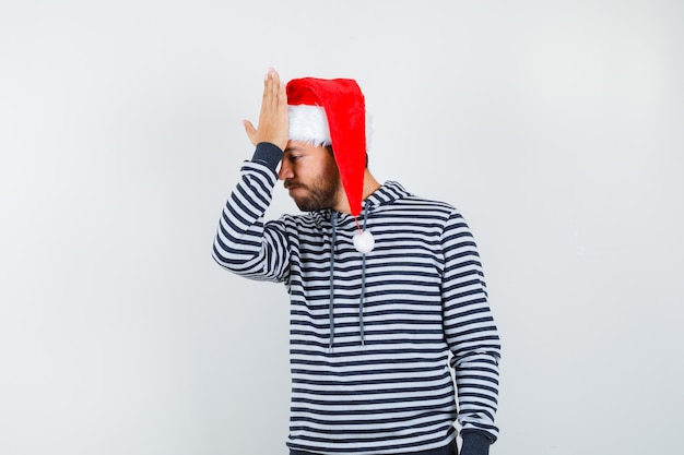 Giovane che tiene la mano sulla fronte in felpa con cappuccio, cappello da babbo natale e sembra smemorato.