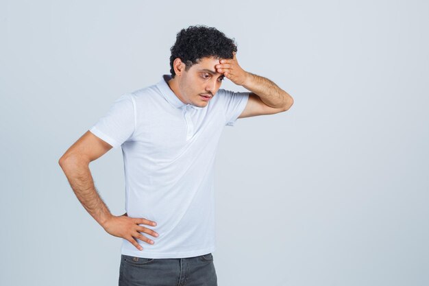 Young man keeping hand on forehead in white t-shirt, pants and looking pensive , front view.