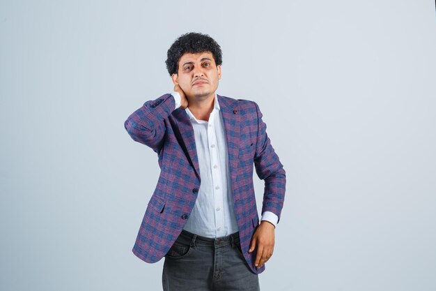 Young man keeping hand on neck in shirt, jacket, pants and looking unwell , front view.