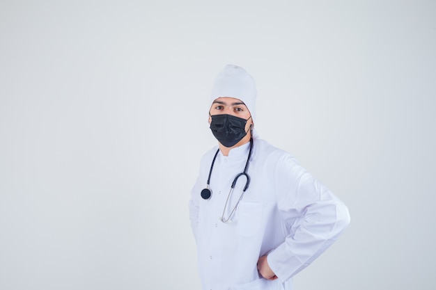 Young man keeping hand on waist in white uniform, mask and looking confident