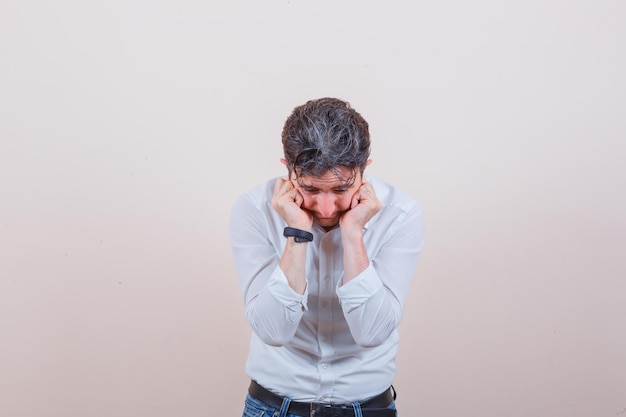 Young man keeping hands on cheeks in white shirt, jeans and looking anxious