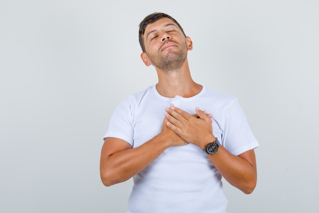 Young man keeping hands on chest in white t-shirt and looking grateful, front view