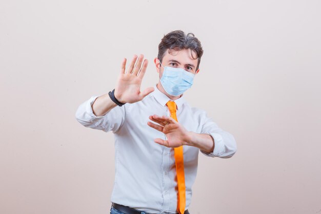 Young man keeping hands to defend himself in shirt, tie and mask and looking anxious