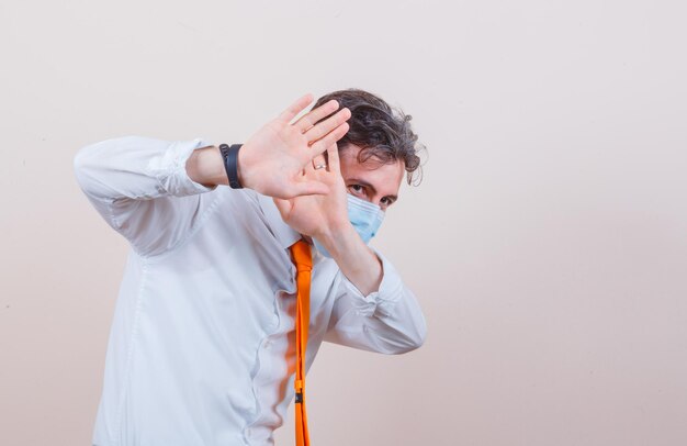 Young man keeping hands to defend himself in shirt, tie, mask and looking frightened