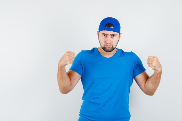 Free photo young man keeping raised fists clenched in blue t-shirt and cap and looking serious