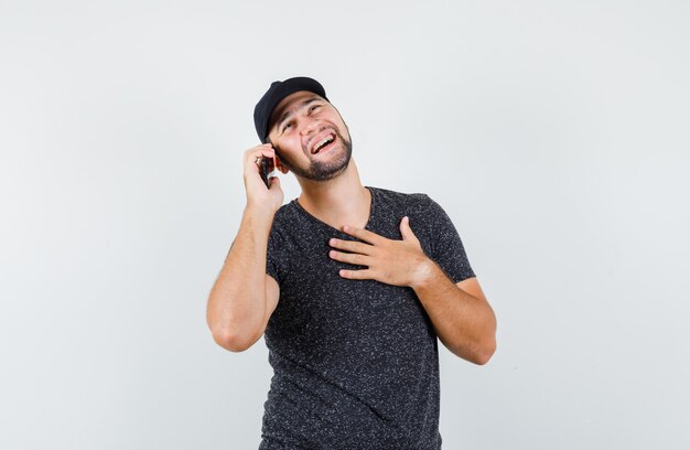 Молодой человек смеется во время разговора по мобильному телефону в футболке и кепке, вид спереди.