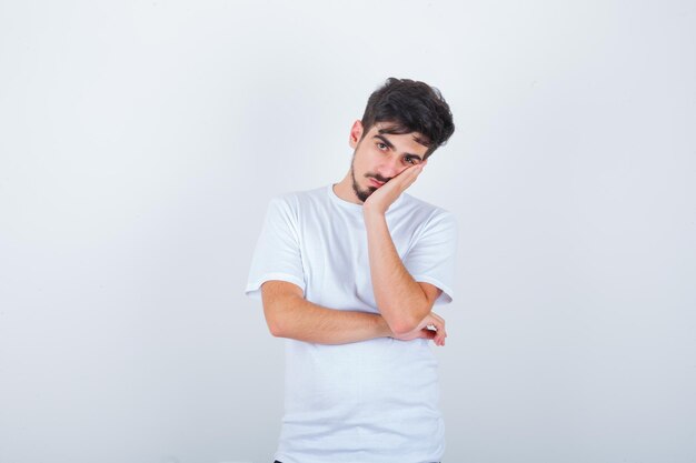 Young man leaning cheek on palm in white t-shirt and looking tired