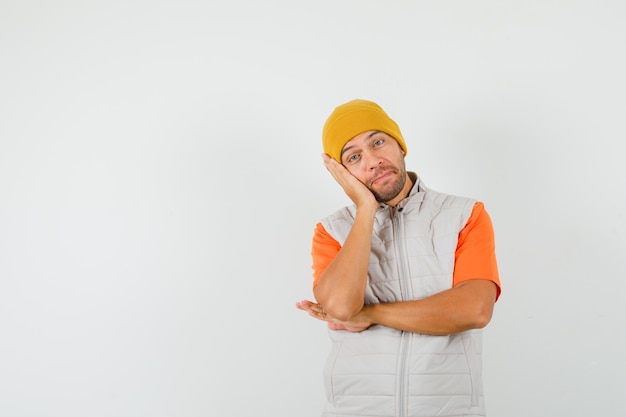 Young man leaning cheek on raised palm in t-shirt, jacket, hat and looking pensive. front view.