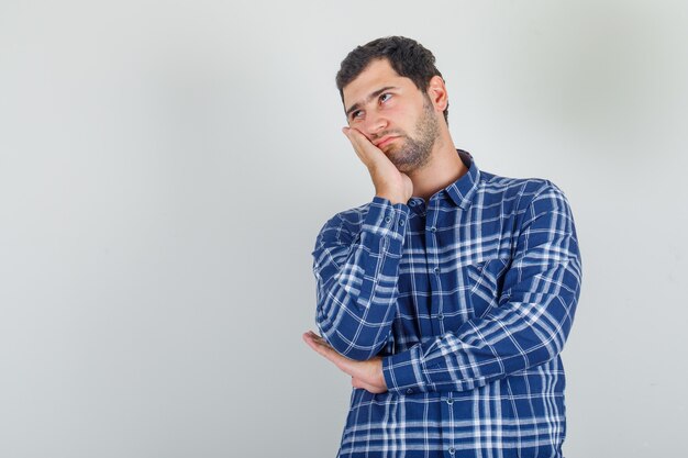 Young man leaning his cheek on raised palm in checked shirt and looking thoughtful