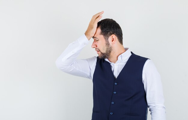 Young man leaning raised hand on forehead in shirt, vest and looking regretful