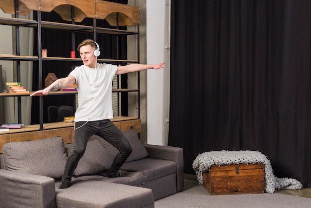 Young man listening music on headphone dancing on sofa at home