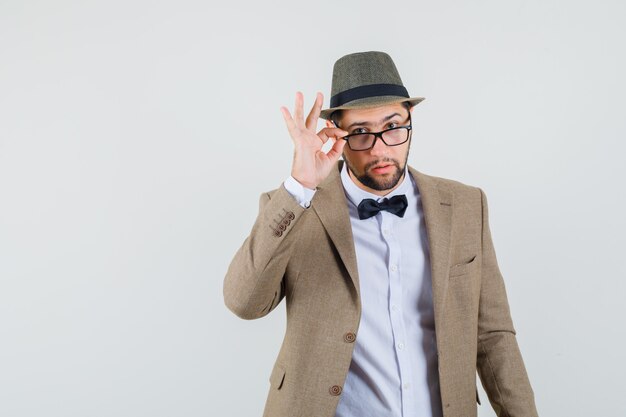 Young man looking attentively over glasses in suit, hat and looking doubtful. front view.