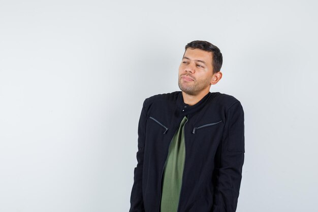 Young man looking away while thinking in t-shirt, jacket and looking hopeful , front view.