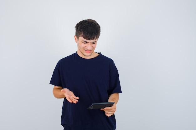 Young man looking at calculator in black t-shirt and looking confused. front view.