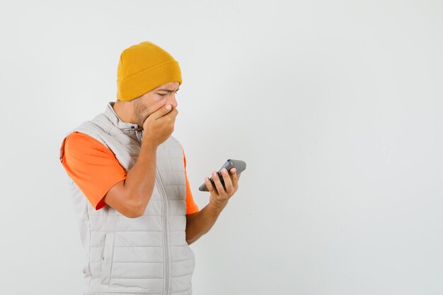 Young man looking at calculator in t-shirt, jacket, hat and looking anxious , front view.