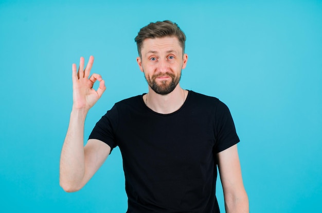 Young man looking at camera by hsowing okay gesture on blue background