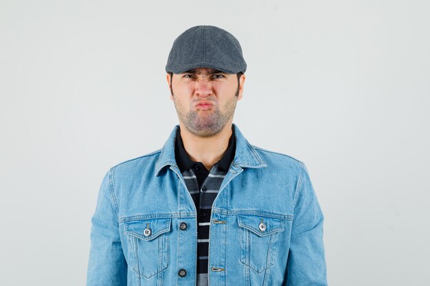 Young man looking at camera in cap, t-shirt, jacket and looking gloomy