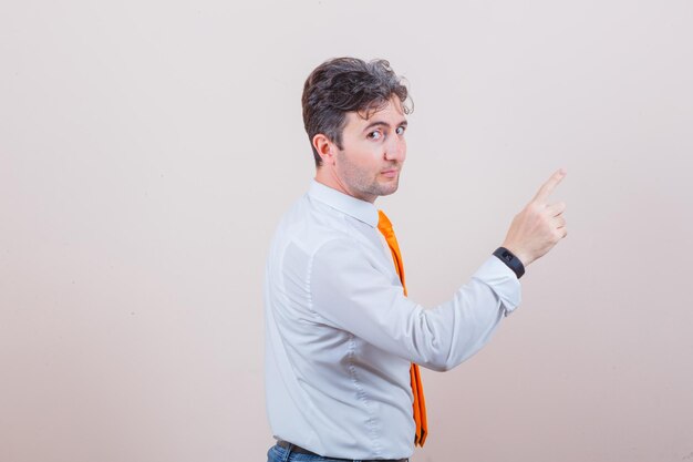 Young man looking at camera, pointing away in white shirt, tie, jeans and looking confident .