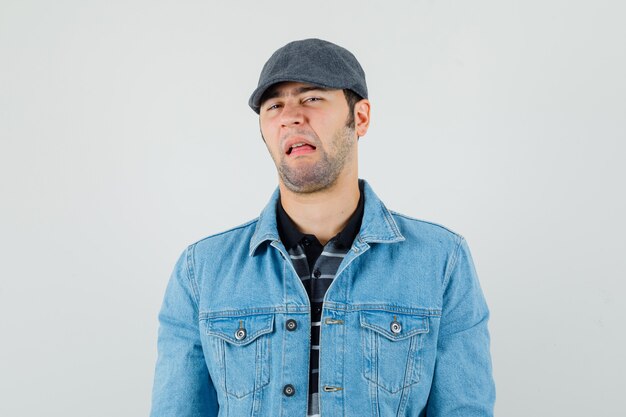 young man looking at camera in scoffing manner in t-shirt, jacket, cap