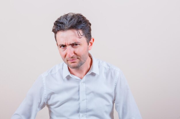 Young man looking at camera while frowning in white shirt and looking suspicious