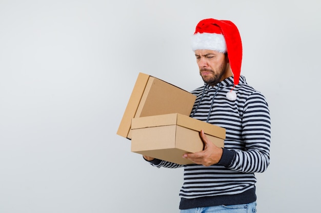 Giovane che guarda scatole di cartone in felpa con cappuccio, cappello di babbo natale e guardando pensieroso, vista frontale.