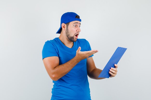 Young man looking over clipboard in blue t-shirt and cap and looking surprised