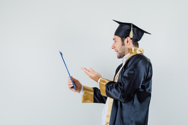 Free photo young man looking at clipboard in graduate uniform and looking confused. .