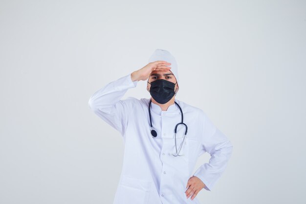 Young man looking far away with hand over head in white uniform, mask and looking focused. front view.