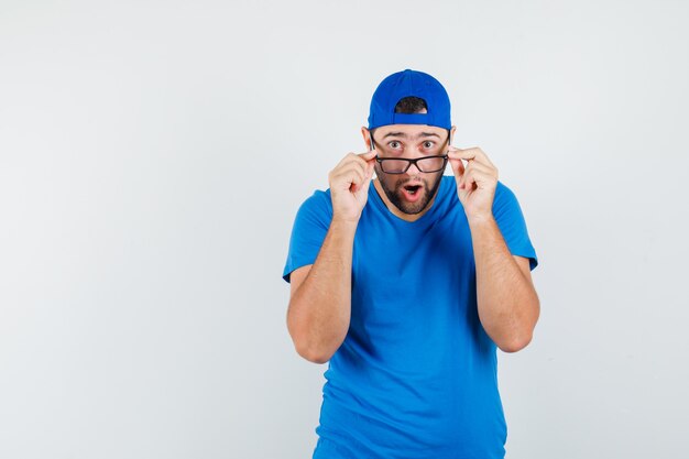 Young man looking over glasses in blue t-shirt and cap and looking shocked