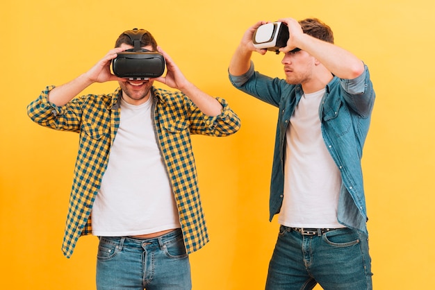 Free photo young man looking at his friend enjoying the virtual reality glasses against yellow backdrop