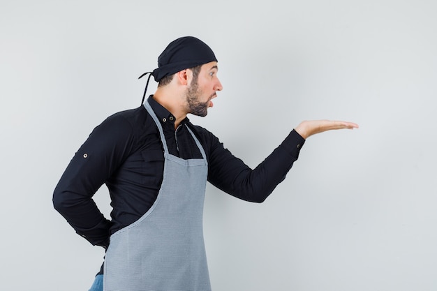 Young man looking at his palm spread aside in shirt, apron and looking wondered , front view.