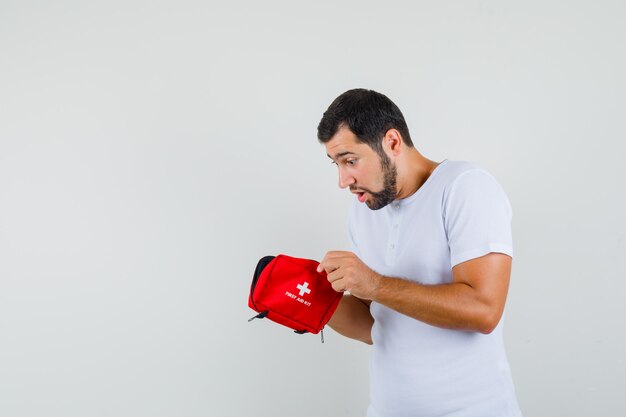 Young man looking into first aid kit in white t-shirt and looking interested. front view. space for text