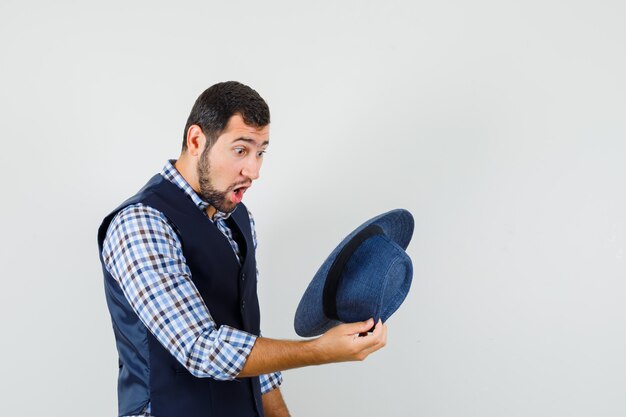 Young man looking into hat in shirt, vest and looking wondered. front view.