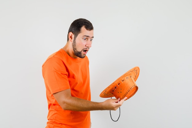 Young man looking into his hat in orange t-shirt and looking wondered , front view.