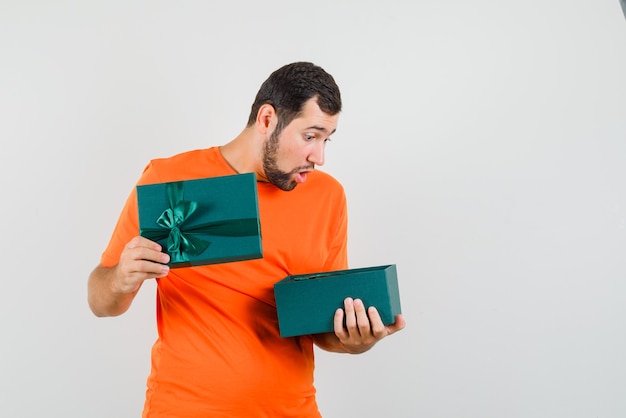 Free photo young man looking into present box in orange t-shirt and looking wondered , front view.