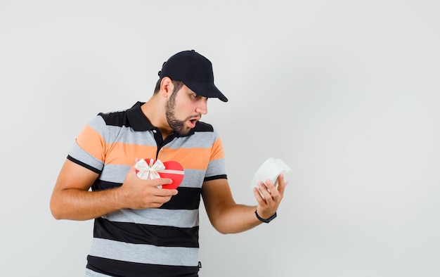 Young man looking into present box in t-shirt, cap and looking shocked , front view.