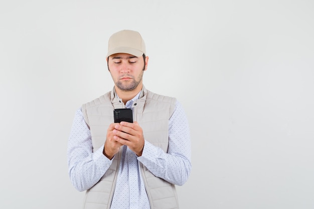 Young man looking at mobile phone in beige jacket and cap and looking focused , front view.