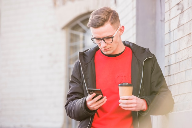 若い男が手に使い捨てのコーヒーカップを保持している携帯電話を見て