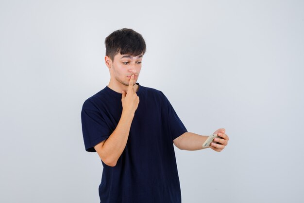Young man looking at mobile phone, keeping finger on lips in black t-shirt and looking puzzled , front view.