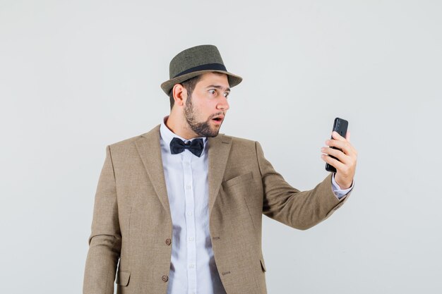 Молодой человек смотрит на мобильный телефон в костюме, шляпе и выглядит потрясенным, вид спереди.
