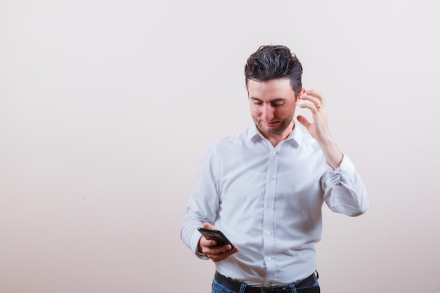 Young man looking at mobile phone while thinking in shirt, jeans and looking irritated