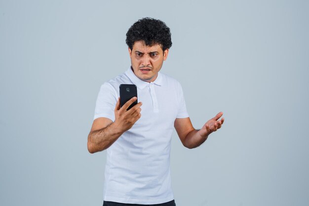 Молодой человек смотрит на мобильный телефон в белой футболке и выглядит смущенным, вид спереди.